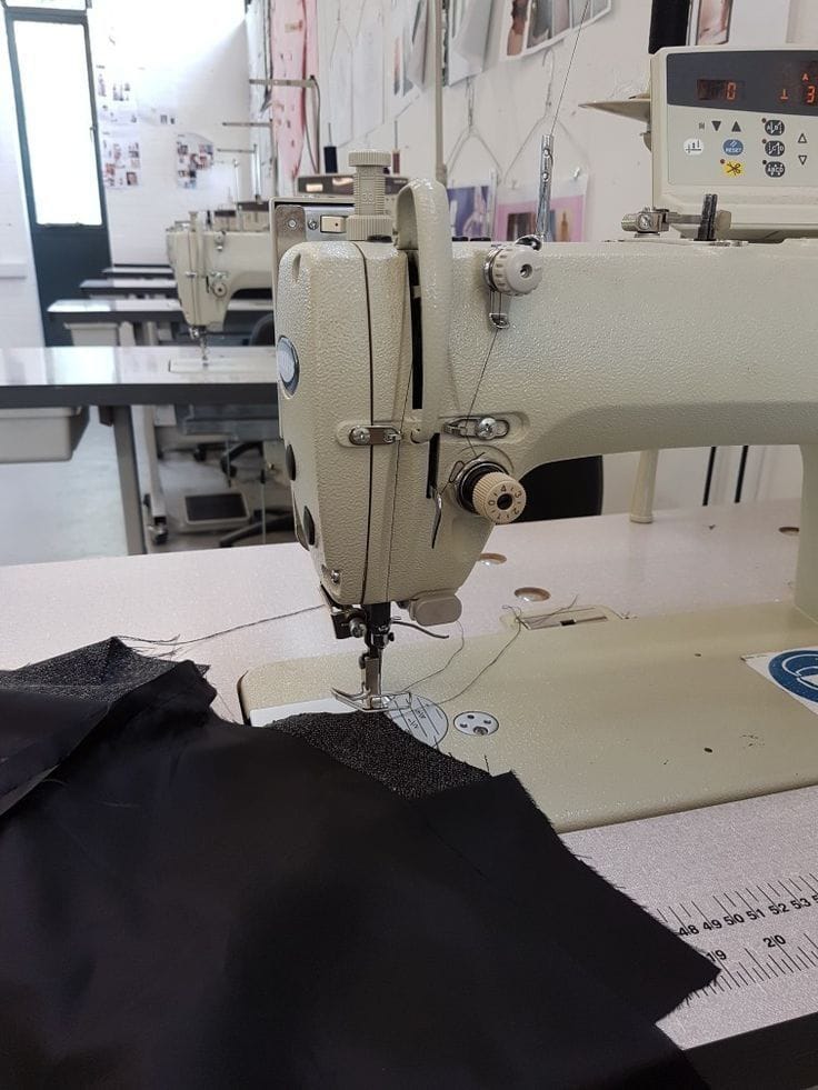 Sewing machine in operation with black fabric on a table in a workshop setting.